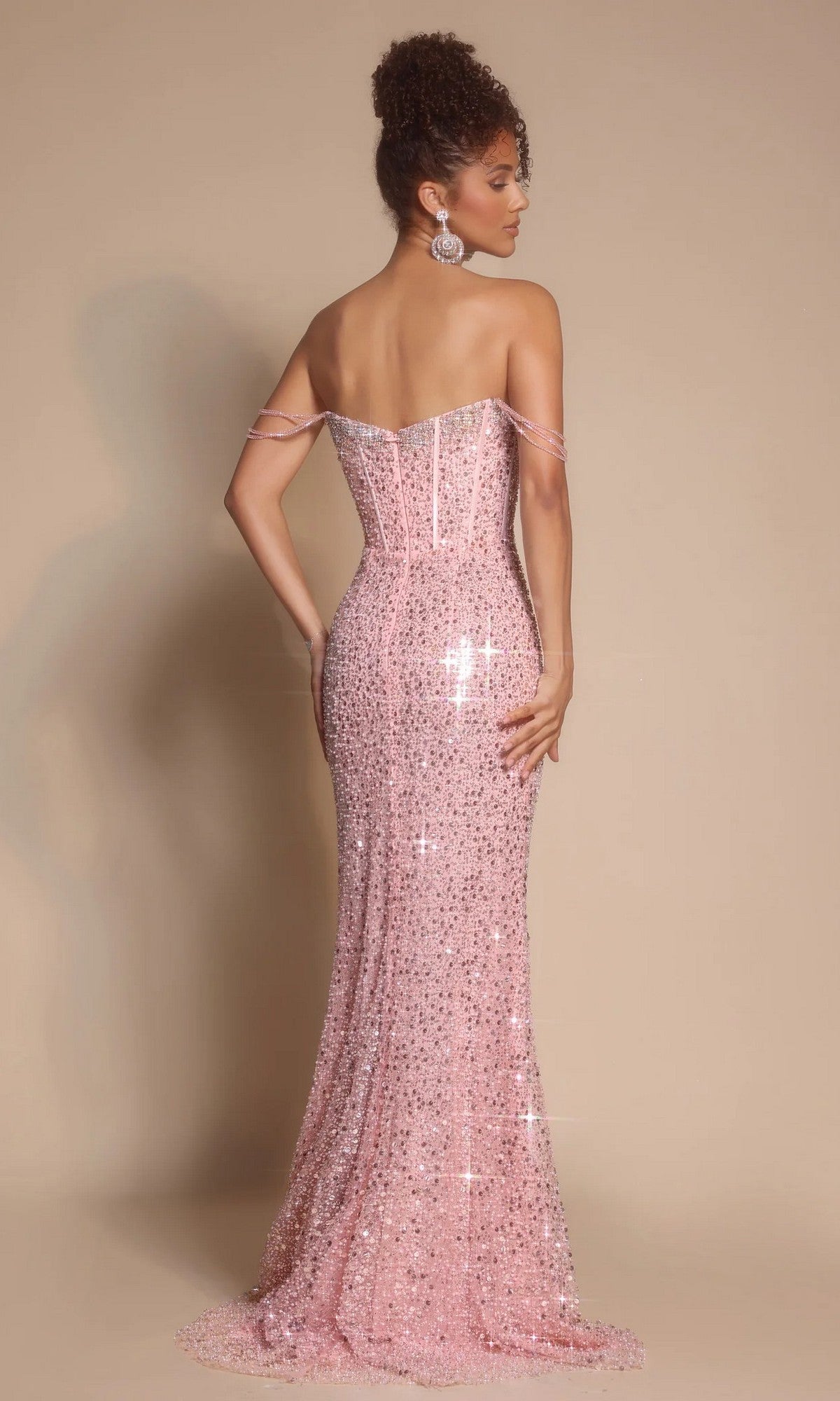 A woman stands with her back to the camera in the Portia and Scarlett XO054 long formal dress, a strapless pink sequin gown featuring a corset bodice, sparkling beaded details, and large earrings against a neutral background.